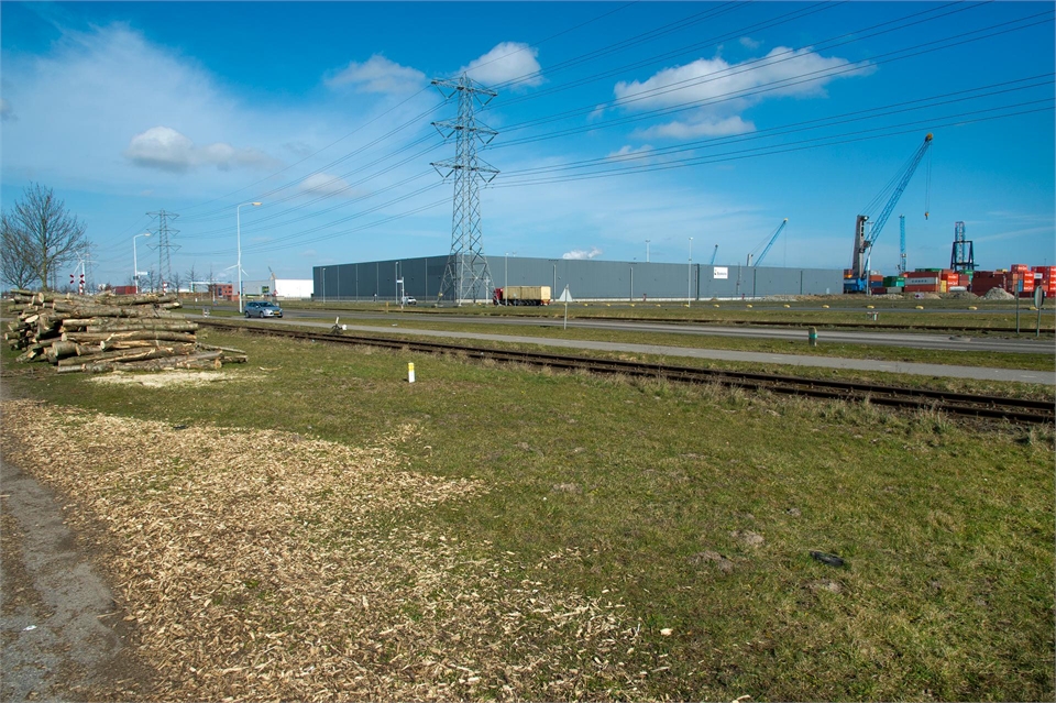 Noord-Brabant - Havengebied Moerdijk: foto van grasveld met in de verte een weg en een groot gebouw en hoogspanningsverbinding.